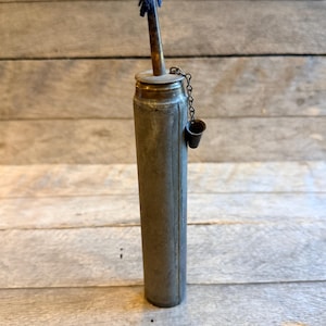 May include: A vintage metal oil lamp with a cylindrical body and a small, attached cup. The lamp has a brass top and a wick with a blue fabric end. The lamp is made of metal and has a rustic, aged appearance.