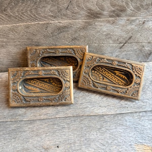 Puede incluir: Tres tiradores de cajón rectangulares de bronce antiguo con intrincados diseños tallados. Cada tirador presenta un hueco ovalado central y patrones florales y geométricos detallados en los bordes. Están dispuestos sobre una superficie de madera envejecida.