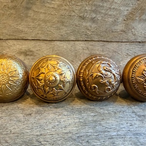May include: Four antique brass doorknobs with detailed embossed patterns. Each knob displays a unique design, including floral and geometric motifs, in a warm, aged gold finish. The knobs are arranged in a row on a weathered wooden surface.