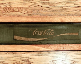May include: A wooden frame with a glass panel displaying the Coca-Cola logo in white text on a dark green background.