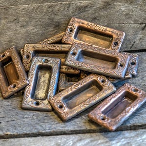 Puede incluir: Un grupo de tiradores de cajones ornamentados, de estilo antiguo, en una variedad de tamaños y formas. Los tiradores están hechos de metal y tienen un acabado desgastado y envejecido.