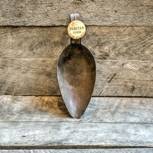 Puede incluir: Una pala de metal oxidada con un mango de madera. El mango tiene una etiqueta redonda amarilla que dice "PURITAN FLOUR 100% PURE".