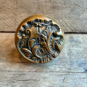 Puede incluir: Un pomo de puerta redondo de latón con un intrincado patrón en relieve de hojas de acanto y volutas. La superficie metálica presenta una pátina pulida y envejecida con áreas oscuras hundidas que resaltan el diseño floral.