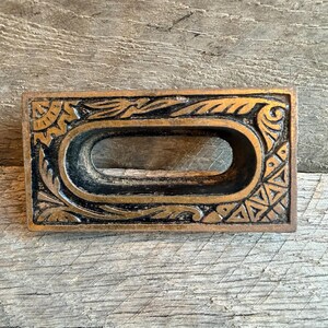 Op de afbeelding: Een rechthoekige bronzen ladegreep met een ovale opening. De greep heeft sierlijke, verhoogde details rond de randen, waaronder bloemen- en geometrische patronen. De achtergrond is een verweerd houten oppervlak.