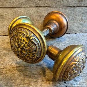 May include: Two antique brass door knobs with ornate floral and geometric designs. The knobs have a warm, aged patina, with a mix of gold and bronze tones. The door knobs are likely vintage and add a touch of classic elegance.