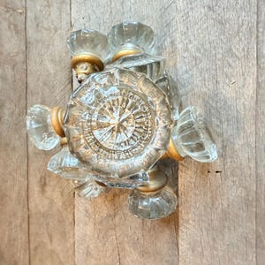 Puede incluir: Un grupo de pomos de puerta de cristal transparente con una base dorada. Los pomos tienen forma facetada y redondeada y están dispuestos sobre una superficie de madera. El pomo central presenta un diseño de estrella.