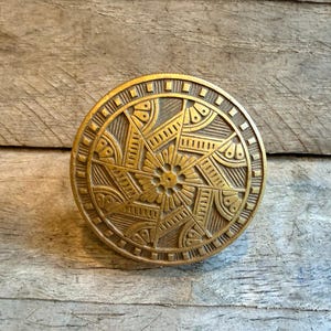 May include: A circular brass knob featuring a detailed geometric pattern with a central flower motif, radiating lines, and small semi-circular accents. The outer edge is bordered by a series of small square indentations, creating a textured finish.