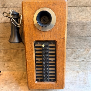 Op de afbeelding: Een vintage wandtelefoon met een rechthoekige eikenhouten behuizing, twee zilverkleurige metalen bellen aan de bovenkant, een ronde zilverkleurige spreekbuis en een zwarte hoorn verbonden met een snoer. Zwart paneel met verticale sleuven en knoppen.