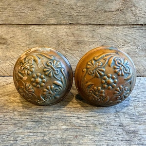 May include: Two antique bronze door knobs with floral and leaf patterns. The knobs have a rounded shape and a weathered appearance, suggesting age and use. The intricate designs are raised and detailed.