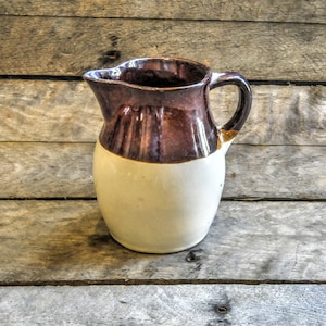 May include: A brown and white ceramic pitcher with a handle. The pitcher has a brown glaze on the top half and a white glaze on the bottom half.