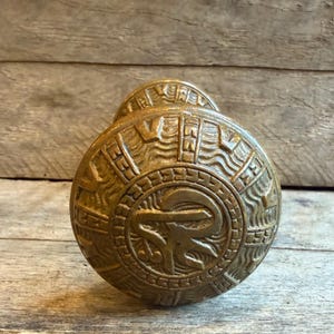 May include: Bronze-colored door knob with an ancient Egyptian design. The knob features a circular pattern with hieroglyphic-style carvings and a central symbol. The background is a weathered wooden surface.