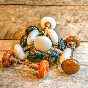 Pode incluir: Uma coleção de puxadores de portas antigos em várias cores e materiais. Os puxadores incluem puxadores de cerâmica e vidro brancos, pretos, verdes e castanhos com hastes de latão ou bronze. Os puxadores estão dispostos sobre uma superfície de madeira.