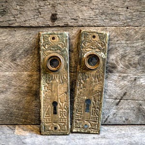 Pode incluir: Duas placas de porta antigas de latão com intrincados desenhos em relevo. Cada placa apresenta uma fechadura, uma abertura circular e padrões ornamentados. As placas retangulares têm um aspeto vintage e desgastado.