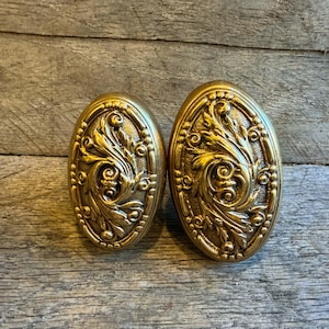 May include: Two antique brass oval door knobs with ornate floral and scrollwork designs. The knobs have a gold finish and are set against a weathered wood background. Decorative hardware.