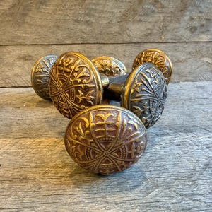 Op de afbeelding: Een verzameling antieke messing deurknoppen met ingewikkelde bloemen- en geometrische patronen. De knoppen hebben een warme, verouderde patina, met tinten goud en brons. De knoppen zijn ovaal van vorm.