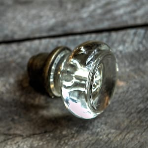 Pode incluir: Uma maçaneta de porta de vidro transparente com uma base de latão. A maçaneta é redonda e tem uma superfície lisa. A base é fixada à maçaneta com um parafuso de metal.