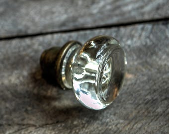 May include: A clear glass door knob with a brass base. The knob is round and has a smooth surface. The base is attached to the knob with a metal screw.
