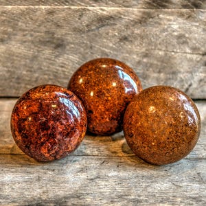 Pode incluir: Três objetos decorativos esféricos polidos, com uma aparência castanho-avermelhada e salpicada. As esferas têm um acabamento brilhante, refletindo a luz, e estão dispostas sobre uma superfície de madeira envelhecida. Os objetos parecem ser de pedra ou material semelhante.