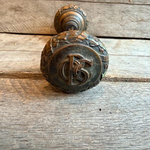 May include: A round, dark bronze-toned metal door knob featuring a raised, intertwined monogram of the letters B and C in the center. The knob is decorated with a relief pattern of laurel leaves around the edge and has a cylindrical base.