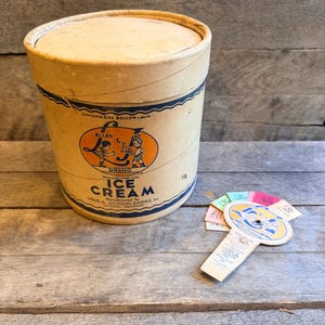 Puede incluir: Contenedor de helado vintage con tapa, con una combinación de colores azul y crema. El contenedor tiene texto que dice "Ice Cream" y "Ellen & Jay Brand". Una pequeña etiqueta circular con muestras de color está junto al contenedor.