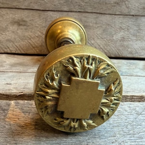 May include: A round brass door knob featuring a central raised square cross motif surrounded by intricate embossed leaf patterns. The metallic surface displays a warm, aged patina with a smooth, rounded neck extending from the back of the knob.