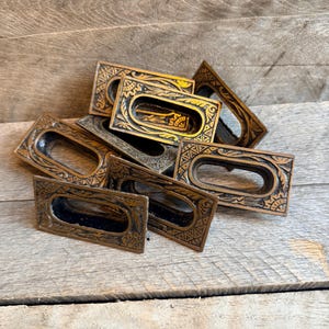 May include: A collection of antique bronze-colored drawer pulls. Each rectangular pull has an oval opening and is decorated with intricate geometric patterns. The pulls are arranged on a weathered wooden surface.