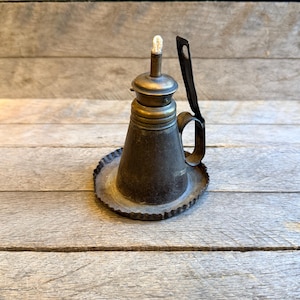 May include: Antique oil lamp with a dark metal finish. The lamp features a conical body, a handle, and a circular base with a decorative edge. A wick is visible at the top. The lamp is designed to sit on a flat surface.