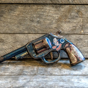 Op de afbeelding: Een blauw speelgoedpistool met een rode en witte sticker waarop "Red Ranger" staat. Het pistool is van metaal en heeft een versleten, vintage uitstraling. Het ligt op een houten oppervlak.