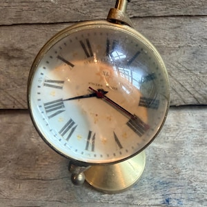 May include: A vintage brass spherical clock with a clear glass cover. The clock face is white with black Roman numerals and black hands. The clock is marked "SWISS MADE".