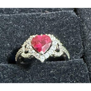 May include: A silver-toned ring featuring a central heart-shaped red gemstone surrounded by a halo of small, clear, round-cut stones. The band and setting are embellished with intricate metalwork and additional clear stones along the shoulders.