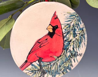 May include: A round, ceramic ornament featuring a vibrant red cardinal perched on a branch with blue-green needles. The bird has a black mask and beak. A red ribbon is attached for hanging. The background is a soft, off-white color.