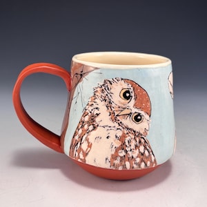 May include: Ceramic mug with a hand-painted owl design. The mug features a light blue background with a brown and white owl illustration. It has a red handle and base, and a cream-colored interior. A unique piece for coffee or tea.