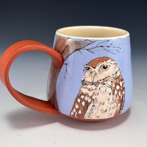 May include: Ceramic mug with a detailed owl illustration. The mug features a light blue background with a brown tree branch design. The handle is a vibrant orange color. The owl has brown and white feathers and yellow eyes.