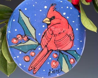 May include: A round, blue ceramic ornament featuring a red cardinal bird perched on a holly branch with red berries. The background is blue with white dots. The ornament is signed "diane".