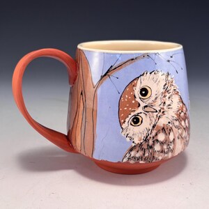 May include: A ceramic mug with a hand-painted owl design. The mug features a light blue background with a brown tree branch and a reddish-orange handle and base. The owl has brown and white feathers and large yellow eyes.