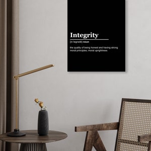 May include: A black and white wall art print with the definition of "Integrity". The word "Integrity" is in a bold white font, with its pronunciation and definition below. The print is displayed in a room with a wooden chair and a side table with a lamp and vase.