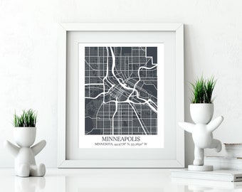 May include: A white framed print of a black and white map of Minneapolis, Minnesota. The map shows the city streets and includes the text "MINNEAPOLIS, MINNESOTA, 44.9778° N, 93.2650° W". The print is displayed on a white surface with two white ceramic planters with green plants.