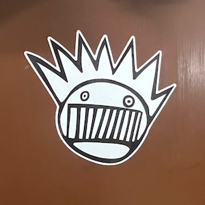 May include: A brown metal cabinet with two doors. The doors have silver handles and a keyhole. A white sticker with a black and white cartoon face is on the left door. The sticker has a spiky crown and a mouth with teeth.