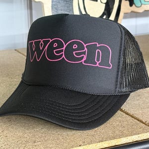 May include: Black trucker hat with a mesh back and a curved brim. The front panel displays the word "ween" in pink, stylized lettering. The hat has a black rope detail above the brim.
