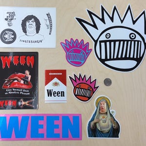 May include: A collection of Ween band stickers and patches, including a black and white sticker with a cartoon face, a pink and blue sticker with a cartoon face, a sticker with a cigarette pack design, a sticker with a woman's face, and a sticker with a band logo. The stickers are on a wooden surface.