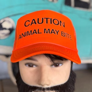 May include: Bright orange trucker hat with the text "CAUTION ANIMAL MAY BITE" in black. The hat has a mesh back and a curved brim. The hat is on a mannequin head with a beard.