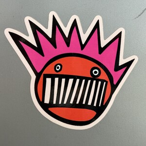 May include: A red and pink sticker with a cartoon face with a black and white striped mouth and two blue eyes. The face has a pink mohawk.