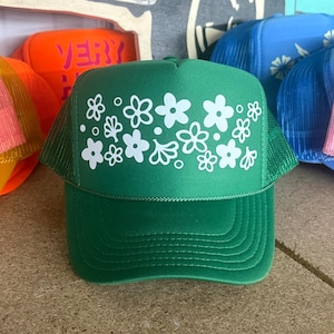 May include: A green trucker hat featuring a white floral pattern on the front foam panel. The hat has a matching green mesh back, a curved brim with stitched detailing, and a structured crown design with a white cord trim above the brim.