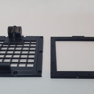 May include: Two black plastic frames, one with a grid pattern and the other with a smooth surface. Both frames are square and have a similar size.