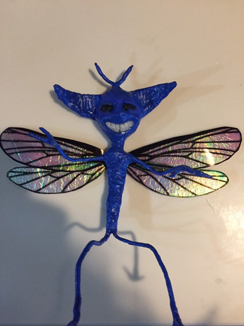 May include: A blue, handmade fairy figurine with iridescent wings. The fairy has a long, thin body with large, pointed ears and a wide smile. The wings are made of a translucent material with a rainbow sheen.