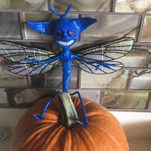 May include: A blue, winged creature with a toothy grin sits atop an orange pumpkin. The creature has a long, thin body and large, iridescent wings.