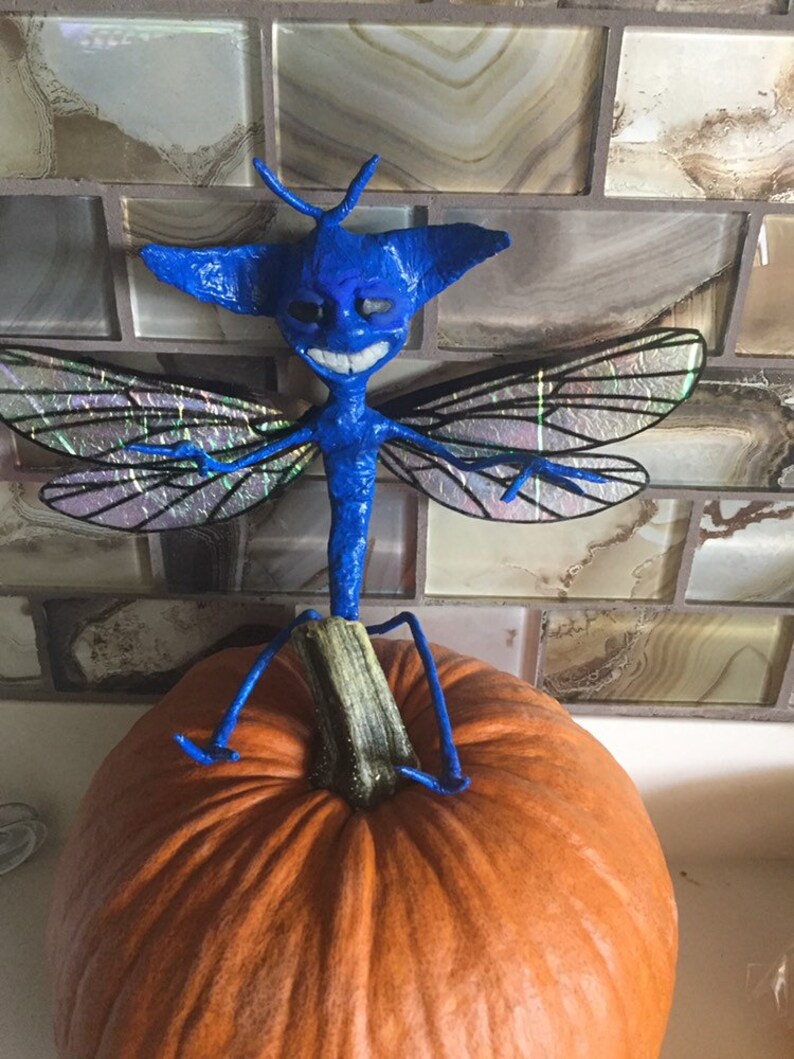 May include: A blue, winged creature with a toothy grin sits atop an orange pumpkin. The creature has a long, thin body and large, iridescent wings.