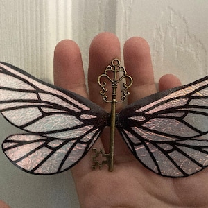 Könnte beinhalten: Ein zarter, schimmernder Schmetterlingsflügel-Anhänger mit einem detaillierten antiken Goldschlüssel-Design. Die Flügel sind durchscheinend mit einem weißen und rosafarbenen, schimmernden Finish.