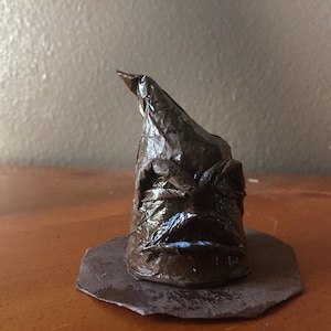 May include: A brown origami Sorting Hat from the Harry Potter series. The hat has a face with a furrowed brow and a serious expression.