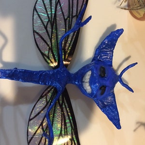 May include: A blue dragonfly sculpture with iridescent wings. The dragonfly has a toothy grin and large, pointed ears.